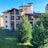 A multi-story residential building with brown roofs and multiple balconies, surrounded by lush green trees and a well-maintained lawn. The sky is clear and blue, contributing to a peaceful atmosphere, and there is a small fenced area in front of the building.