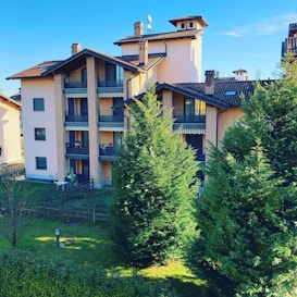A multi-story residential building with brown roofs and multiple balconies, surrounded by lush green trees and a well-maintained lawn. The sky is clear and blue, contributing to a peaceful atmosphere, and there is a small fenced area in front of the building.