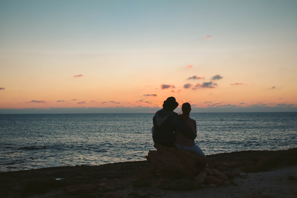 silhouette of couple sitting on rock at shore during golden hour