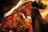 An intense, vivid close-up of a burning fire with logs and flames. The image captures the dynamic movement of the flames enveloping the charred wood, highlighting the intricate textures of the burning logs and ashes.