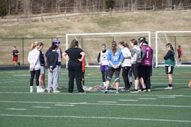 A group of people gathers on a sports field, likely for a team discussion or practice session. They are dressed in athletic clothing, and some are holding or standing near lacrosse sticks. The setting appears to be an outdoor sports complex with a netted goal visible in the background. The field markings indicate it is a standard sports field, and the weather looks cool as some individuals are wearing long sleeves.