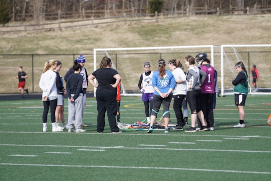 A group of people gathers on a sports field, likely for a team discussion or practice session. They are dressed in athletic clothing, and some are holding or standing near lacrosse sticks. The setting appears to be an outdoor sports complex with a netted goal visible in the background. The field markings indicate it is a standard sports field, and the weather looks cool as some individuals are wearing long sleeves.