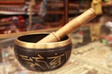 A Tibetan singing bowl made of metal with intricate golden inscriptions, paired with a wooden mallet resting on the rim. The background appears to be a shop display with various colorful fabrics and items, slightly blurred.