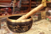 A Tibetan singing bowl made of metal with intricate golden inscriptions, paired with a wooden mallet resting on the rim. The background appears to be a shop display with various colorful fabrics and items, slightly blurred.