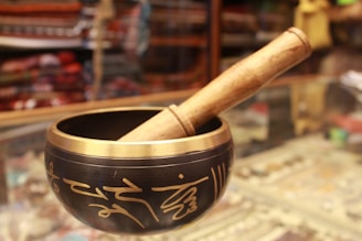A Tibetan singing bowl made of metal with intricate golden inscriptions, paired with a wooden mallet resting on the rim. The background appears to be a shop display with various colorful fabrics and items, slightly blurred.