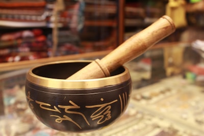 A Tibetan singing bowl made of metal with intricate golden inscriptions, paired with a wooden mallet resting on the rim. The background appears to be a shop display with various colorful fabrics and items, slightly blurred.