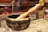A Tibetan singing bowl made of metal with intricate golden inscriptions, paired with a wooden mallet resting on the rim. The background appears to be a shop display with various colorful fabrics and items, slightly blurred.