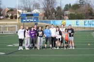 A group of people stand together on a sports field, each holding a lacrosse stick. They are dressed casually, suggesting they are gathered for a practice or informal game. Behind them, a scoreboard and bleachers are visible, with a mural on the wall depicting a Spartan helmet and the word 'LEGENDS.' Trees and houses can be seen in the background.