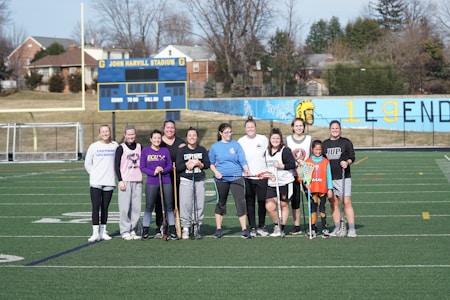 A group of people stand together on a sports field, each holding a lacrosse stick. They are dressed casually, suggesting they are gathered for a practice or informal game. Behind them, a scoreboard and bleachers are visible, with a mural on the wall depicting a Spartan helmet and the word 'LEGENDS.' Trees and houses can be seen in the background.