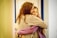 Two individuals embrace in a hug, showing close emotional connection. Both are dressed in casual clothing, one wearing a beige sweater and the other in a purple one. The background is softly lit with yellow tones, likely indoors.
