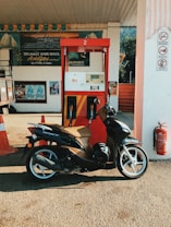 A motorcycle is parked in front of a gas pump at a petrol station. The pump is red and features signage with decals and pricing information. Surrounding the station are orange safety cones, various signs on the walls, and promotional banners hanging. The setting appears to be outdoors with daylight illuminating the area.