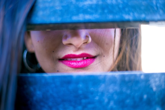 A close-up of a woman's face, focusing on her lips and nose. Her lips are painted bright pink, and she has a gold nose ring. The view is partially obscured by blue horizontal bars.