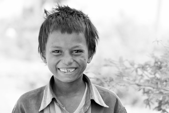 A young boy with tousled hair is smiling broadly at the camera. His expression is joyful, and he appears to be in a natural, outdoor setting with blurred foliage in the background. The photograph is in black and white, emphasizing the textures and contrasts.
