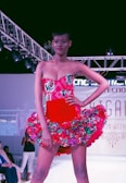 A model confidently walks down a runway wearing a vibrant red dress adorned with floral patterns. The dress features a strapless top and a flared skirt accentuated with multicolored flowers. Overhead lighting highlights the model and creates a dramatic atmosphere on the fashion show stage.