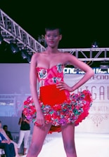 A model confidently walks down a runway wearing a vibrant red dress adorned with floral patterns. The dress features a strapless top and a flared skirt accentuated with multicolored flowers. Overhead lighting highlights the model and creates a dramatic atmosphere on the fashion show stage.