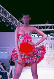 A model confidently walks down a runway wearing a vibrant red dress adorned with floral patterns. The dress features a strapless top and a flared skirt accentuated with multicolored flowers. Overhead lighting highlights the model and creates a dramatic atmosphere on the fashion show stage.