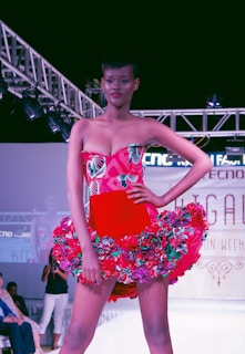 A model confidently walks down a runway wearing a vibrant red dress adorned with floral patterns. The dress features a strapless top and a flared skirt accentuated with multicolored flowers. Overhead lighting highlights the model and creates a dramatic atmosphere on the fashion show stage.