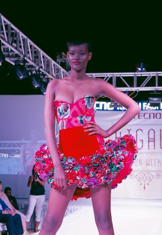 A model confidently walks down a runway wearing a vibrant red dress adorned with floral patterns. The dress features a strapless top and a flared skirt accentuated with multicolored flowers. Overhead lighting highlights the model and creates a dramatic atmosphere on the fashion show stage.