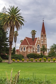 A picturesque brick church with red-tiled roofs and a tall, pointed steeple topped with a cross is surrounded by lush greenery and tall palm trees. The scene is set on a well-manicured lawn under an overcast sky, creating a tranquil and serene atmosphere.