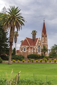 A picturesque brick church with red-tiled roofs and a tall, pointed steeple topped with a cross is surrounded by lush greenery and tall palm trees. The scene is set on a well-manicured lawn under an overcast sky, creating a tranquil and serene atmosphere.