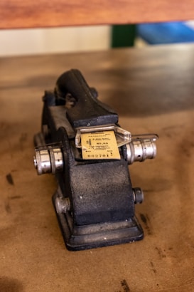 An old, industrial-style machine is placed on a wooden surface. It appears to be a numbering or ticket machine, with a handle on top for manual operation. The machine is made of metal and has a worn, vintage appearance. A ticket is visible at the front, with typography printed on it.