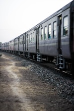 A long train with several passenger cars moves along a track bordered by gravel and a dirt path. The railway cars are dark in color and the scene seems to be captured in motion on a somewhat overcast day.