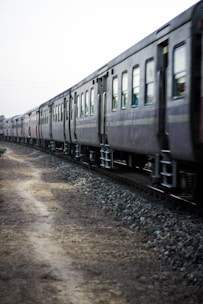 A long train with several passenger cars moves along a track bordered by gravel and a dirt path. The railway cars are dark in color and the scene seems to be captured in motion on a somewhat overcast day.
