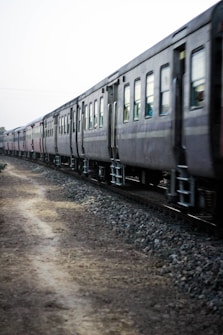 A long train with several passenger cars moves along a track bordered by gravel and a dirt path. The railway cars are dark in color and the scene seems to be captured in motion on a somewhat overcast day.