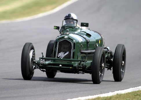A vintage racing car, likely from the early to mid-20th century, is driving on a racetrack. It features a dark green color, large exposed wheels, and a classic design with minimal bodywork. The driver is wearing a helmet and is partially visible. The background consists of a blurred view of the race track, indicating motion.