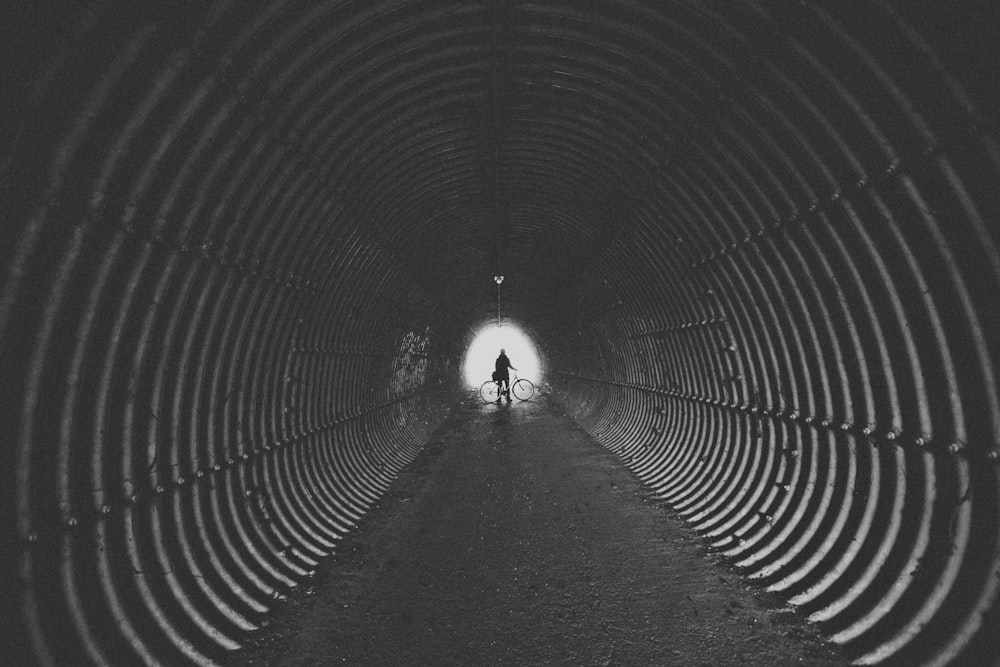 Foto en escala de grises de un hombre sosteniendo su bicicleta en un túnel