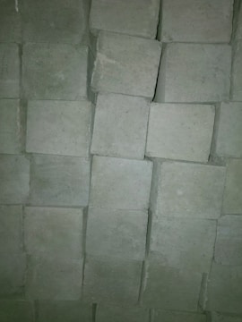 A close-up view of a stack of grayish cement blocks arranged in an orderly fashion. The blocks have a rough texture and are slightly irregular in shape, creating a pattern through their arrangement.