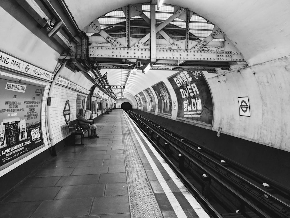 grayscale photo of man sitting on metal bench waiting for train