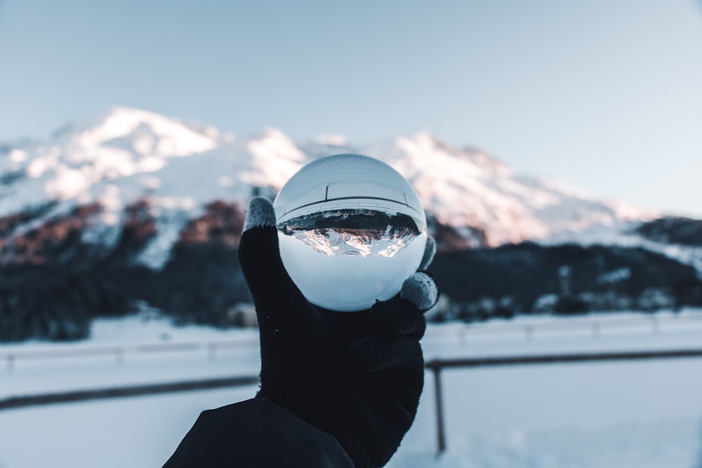 person holding glass crystal ball below snow-covered mountains during daytime