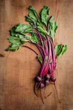 A bunch of fresh beetroots with vibrant green leaves and deep red-purple stems and roots lie on a wooden surface. The wood displays a light brown color with visible grain patterns.