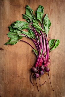 A bunch of fresh beetroots with vibrant green leaves and deep red-purple stems and roots lie on a wooden surface. The wood displays a light brown color with visible grain patterns.