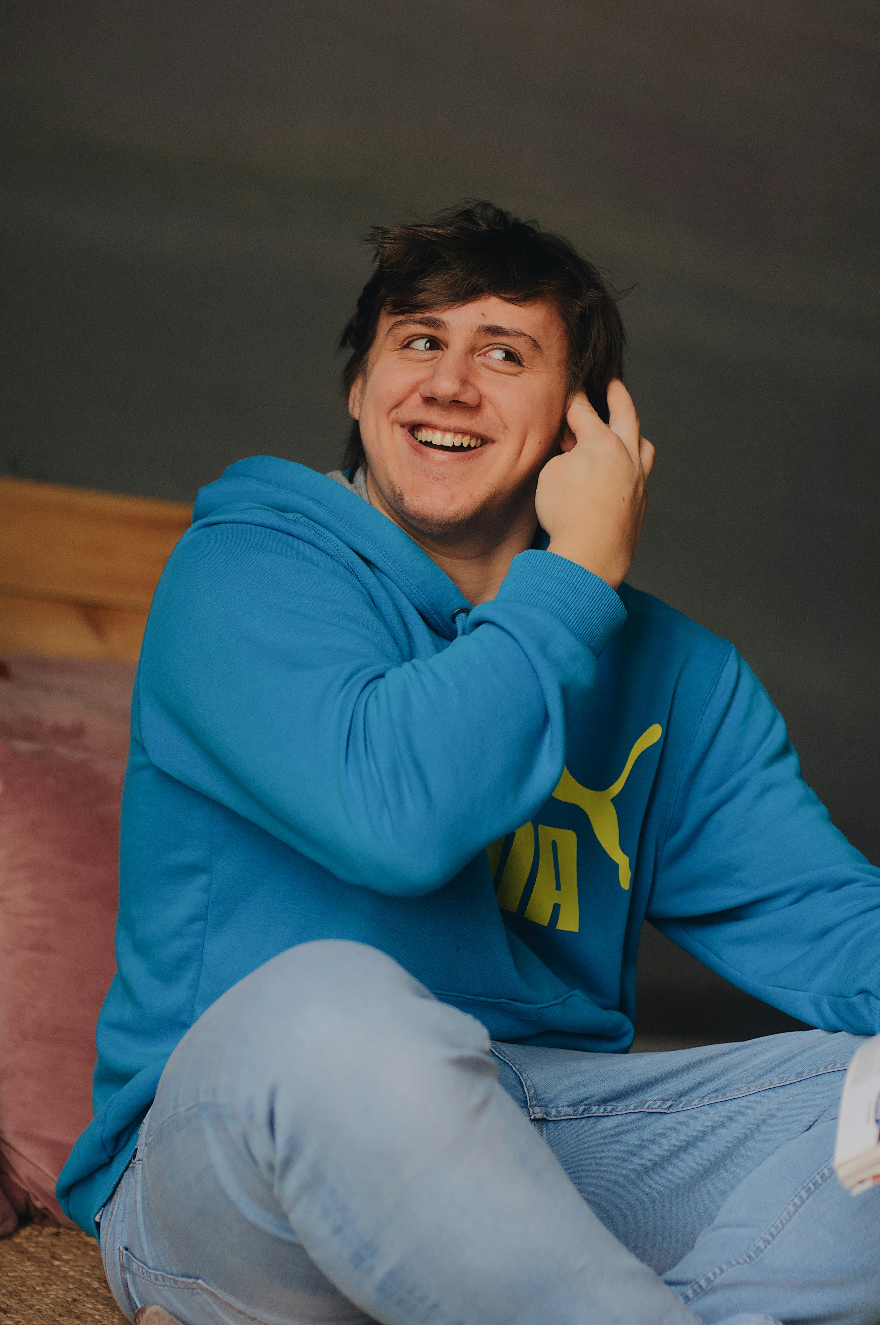 pria tersenyum dengan hoodie pullover PUMA biru duduk di tempat tidur coklat
