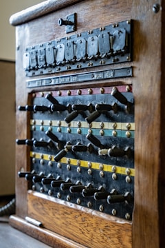 An antique wooden telephone switchboard with rows of levers and switches, characterized by aged brass accents and visible labels indicating various connections. The switchboard features different colored stripes, including blue, red, and yellow, that run horizontally along the panel, contributing to its vintage appearance.