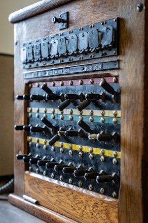 An antique wooden telephone switchboard with rows of levers and switches, characterized by aged brass accents and visible labels indicating various connections. The switchboard features different colored stripes, including blue, red, and yellow, that run horizontally along the panel, contributing to its vintage appearance.
