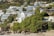 The image features a scenic coastal hillside with a series of modern, multi-story homes and villas nestled among lush green foliage. The architecture is sleek and contemporary with large glass windows and balconies that offer a view of the ocean. The hillside is densely packed with trees and shrubs, creating a verdant backdrop. At the forefront, there is a rocky beach with sand and a stone wall separating the properties from the shoreline.