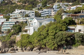 The image features a scenic coastal hillside with a series of modern, multi-story homes and villas nestled among lush green foliage. The architecture is sleek and contemporary with large glass windows and balconies that offer a view of the ocean. The hillside is densely packed with trees and shrubs, creating a verdant backdrop. At the forefront, there is a rocky beach with sand and a stone wall separating the properties from the shoreline.