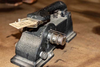 A vintage mechanical device with a sturdy build, featuring metal dials and a ticket inserted in a clamp. The surface appears textured and the background is blurred.