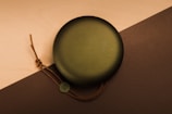 A circular portable speaker with a perforated grill and brown leather strap lies on a two-toned surface, featuring light wood and dark brown colors. The speaker has a small monogrammed tag attached to the strap.