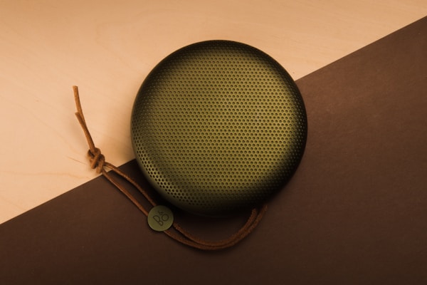 A circular portable speaker with a perforated grill and brown leather strap lies on a two-toned surface, featuring light wood and dark brown colors. The speaker has a small monogrammed tag attached to the strap.