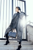 A person stands confidently on a set of stone steps, wearing a stylish outfit that includes a dark headscarf and a patterned long coat. They hold a black designer handbag and wear elegant black high-heeled boots. The background features vertical metal railings with bright sunlight streaming through.
