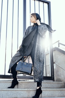 woman in gray coat holding black handbag standing on gray stairs during daytime