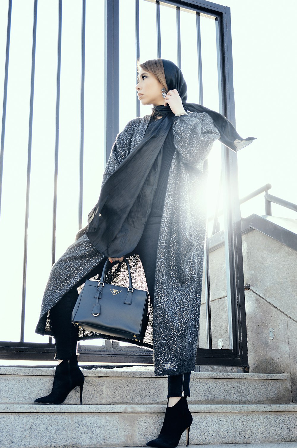 Femme en manteau gris tenant un sac à main noir debout sur des escaliers gris pendant la journée