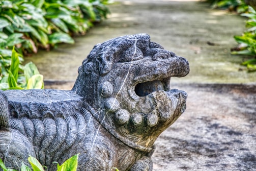 A detailed stone sculpture of a mythical creature, resembling a lion or dragon, set in an outdoor environment. Lush green foliage surrounds the sculpture, adding to the natural setting.
