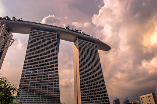 Two tall, modern towers are connected by a rooftop structure with greenery on top, set against a dramatic sky filled with large, billowing clouds. The architectural design is striking and innovative.