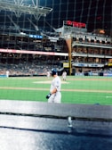 A baseball game scene at a stadium during nighttime, with players on the field and a batter in focus wearing a white uniform. The stadium's stands are partially filled, and several advertisements and brand logos are visible, including Toyota and Western Metal Supply Co. The netting in front of the camera indicates the perspective is from behind home plate.