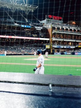 A baseball game scene at a stadium during nighttime, with players on the field and a batter in focus wearing a white uniform. The stadium's stands are partially filled, and several advertisements and brand logos are visible, including Toyota and Western Metal Supply Co. The netting in front of the camera indicates the perspective is from behind home plate.